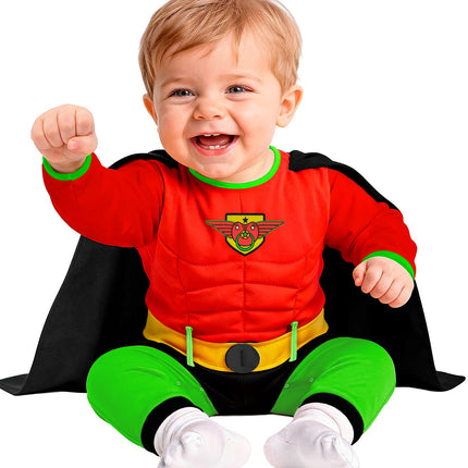 Baby in rood superheldenpak met geel schildlogo en speen, zittend met zwarte cape