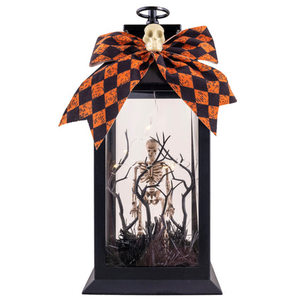 Halloween lantaarn zwart met skelet tussen kale takken, lichtjes en oranje-zwarte strik met schedeltje